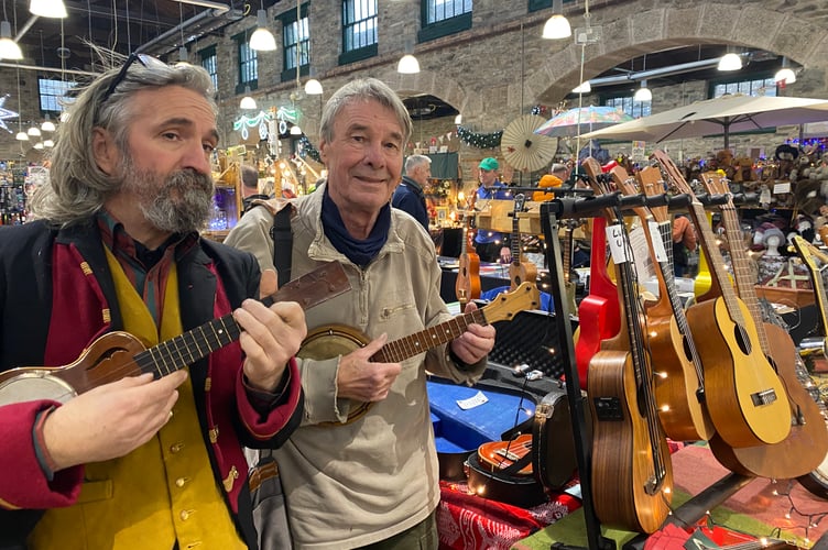 Ukulele player Neil Campbell serenading shoppers at Tavistock Pannier Market during late night opening with customer John Spence, customer and silversmith.