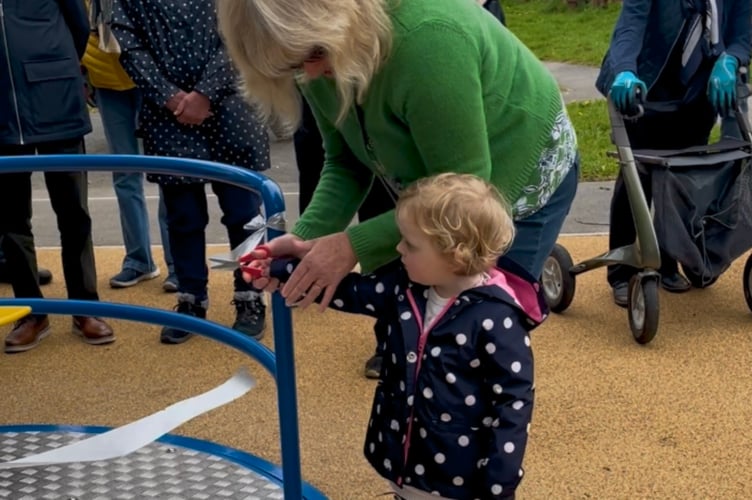 Young Imogen McFarlane cutting the Yelverton Play Park ceremonial ribbon on the new roundabout, with a little help.